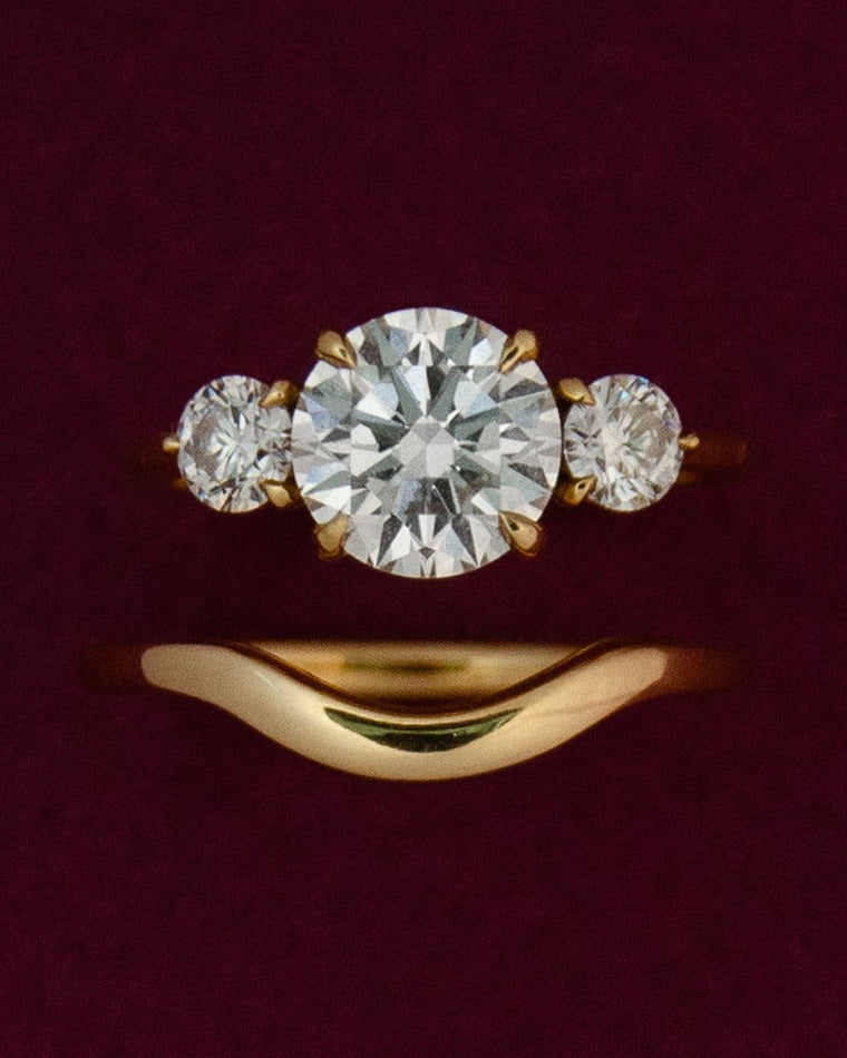 Gold curved wedding band with a 3 stone engagement ring on a red background